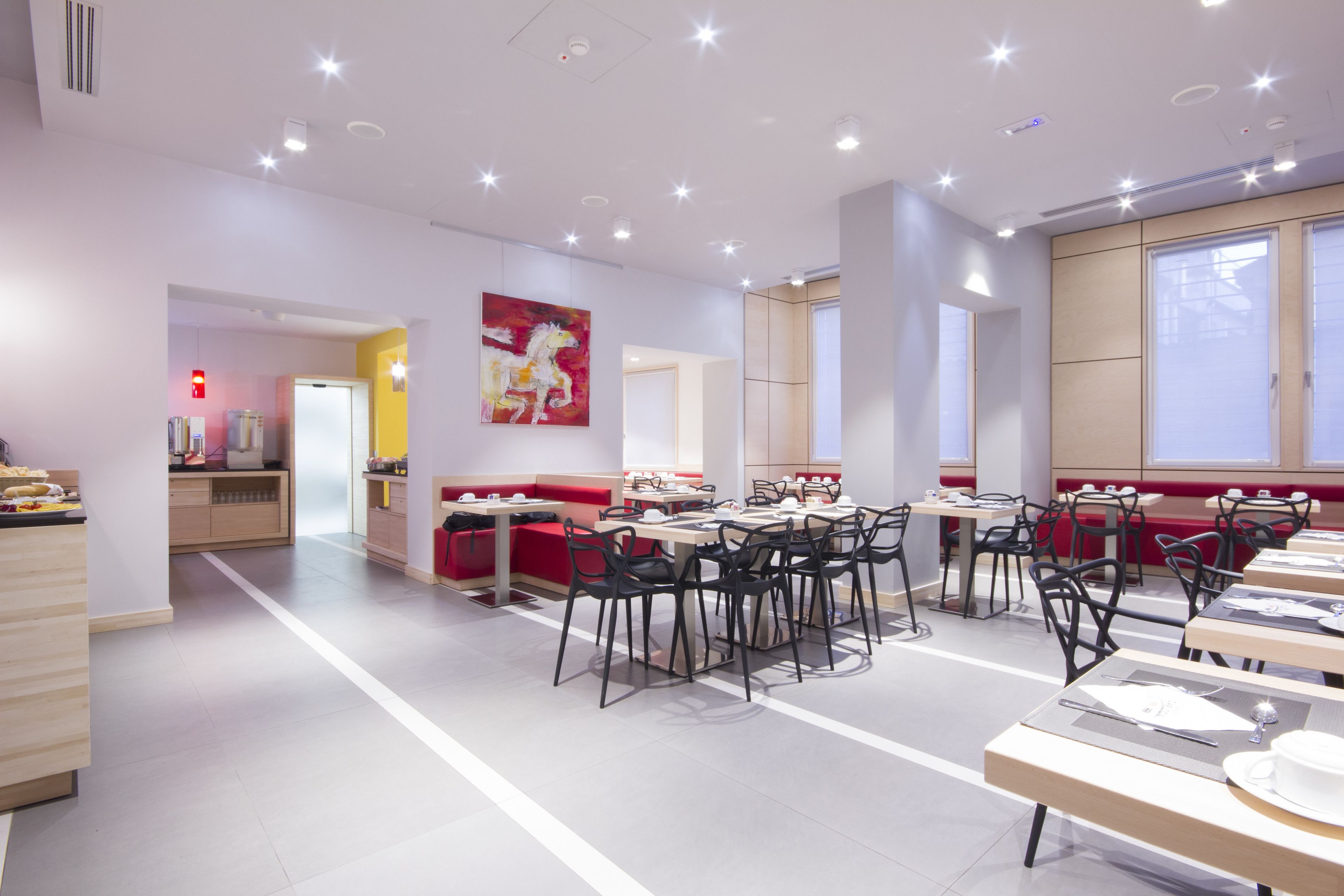 Un espace repas moderne avec plusieurs tables et chaises, un coin salon rouge et une grande fenêtre laissant entrer la lumière naturelle.