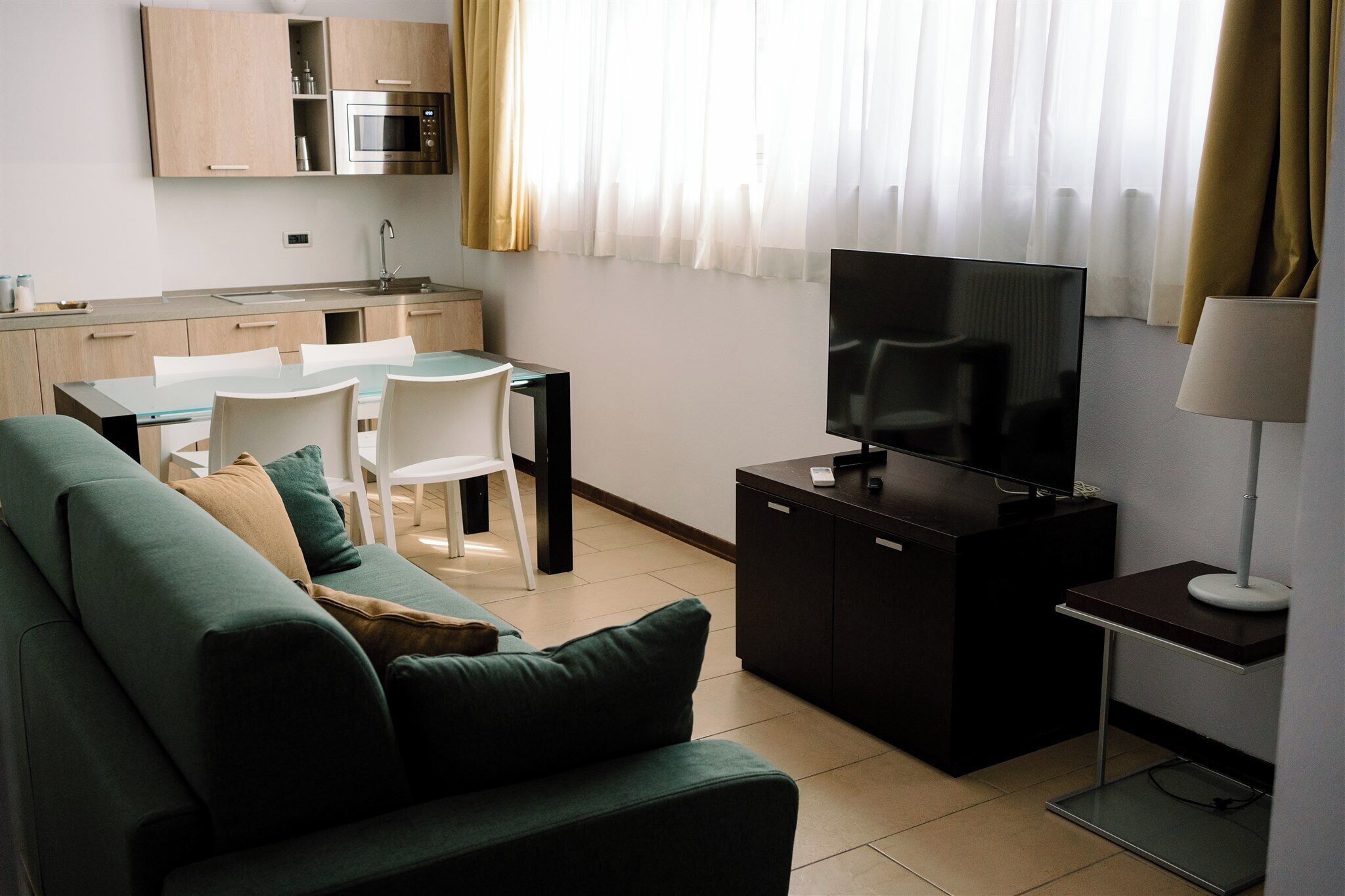 A modern living room with a green sofa, a dining table with chairs, a TV on a cabinet, and a kitchen area with cabinets and a microwave.