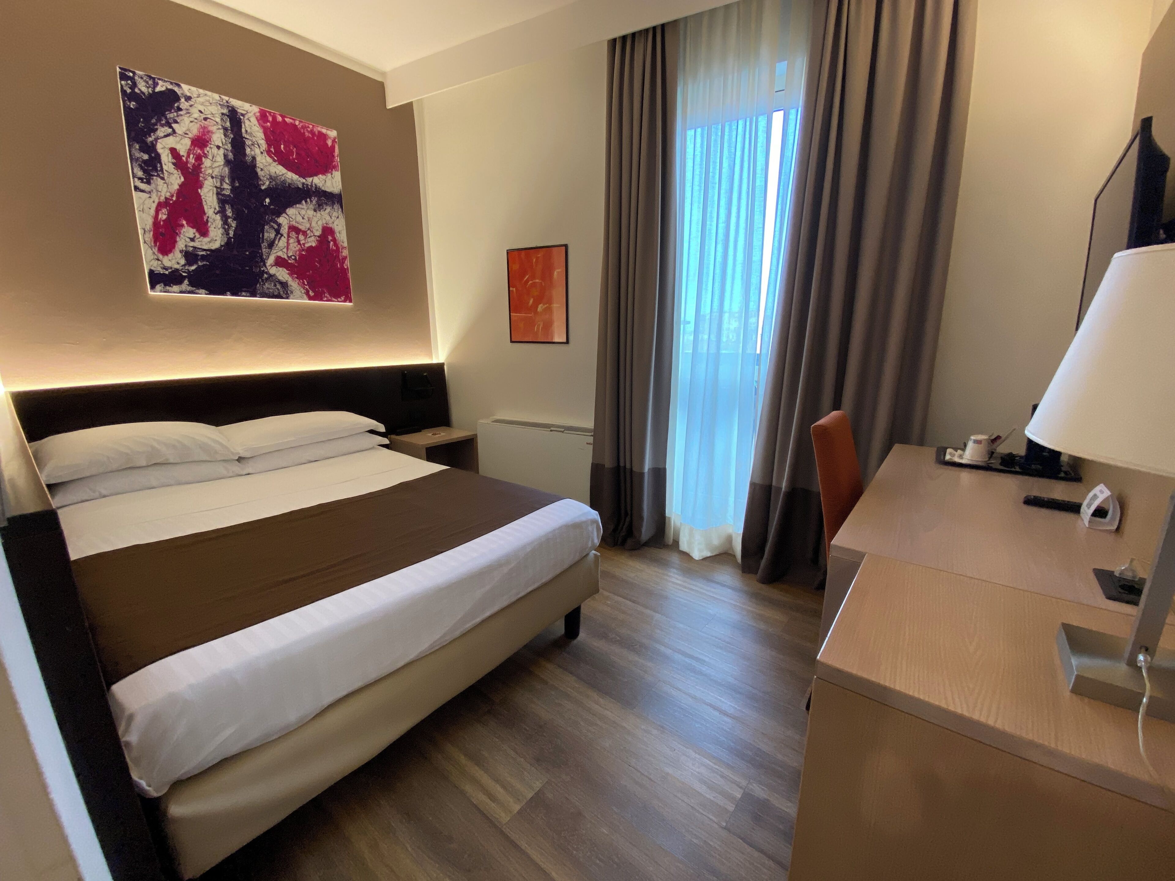 Habitación de hotel con una cama grande, un escritorio con lámpara, un cuadro en la pared y una ventana con cortinas.