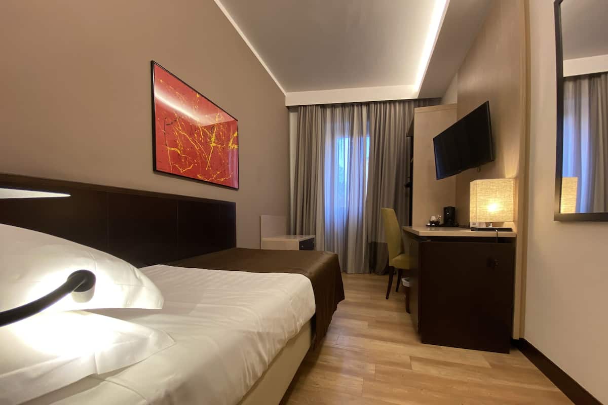 Habitación de hotel con cama, escritorio, silla, televisor y ventana con cortinas.