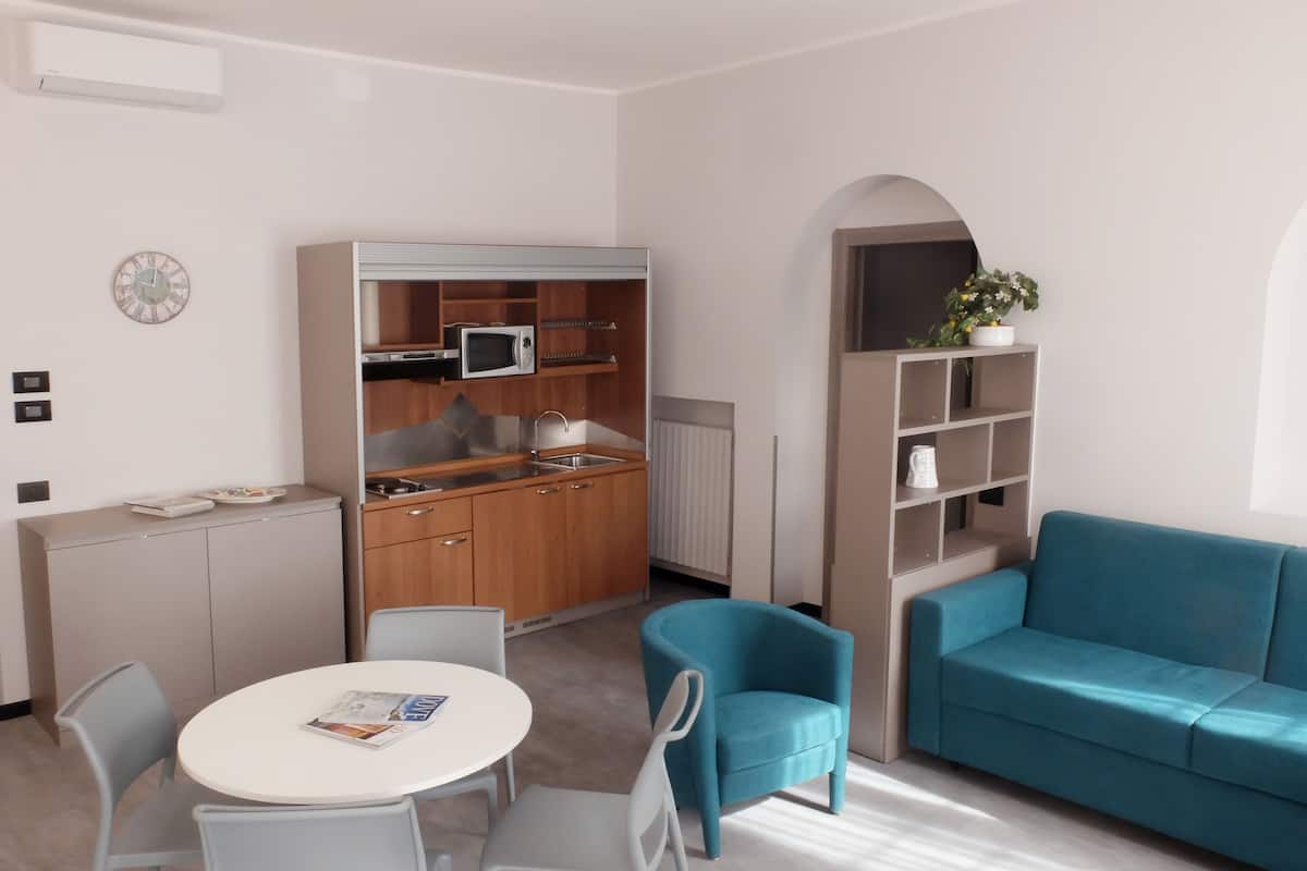 Un espacio habitable compacto con kitchenette, un área de comedor con mesa y sillas redondas, un sofá color turquesa y una pequeña estantería.