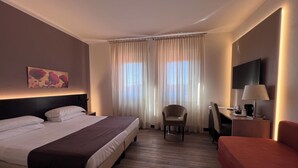A hotel room with a bed, a chair, a desk with a lamp, and a TV on the wall. - Hotel Carlton (Ferrara)