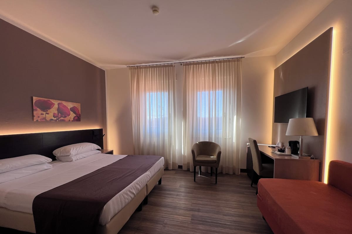 Habitación de hotel con cama, una silla, escritorio con lámpara y televisión en la pared.