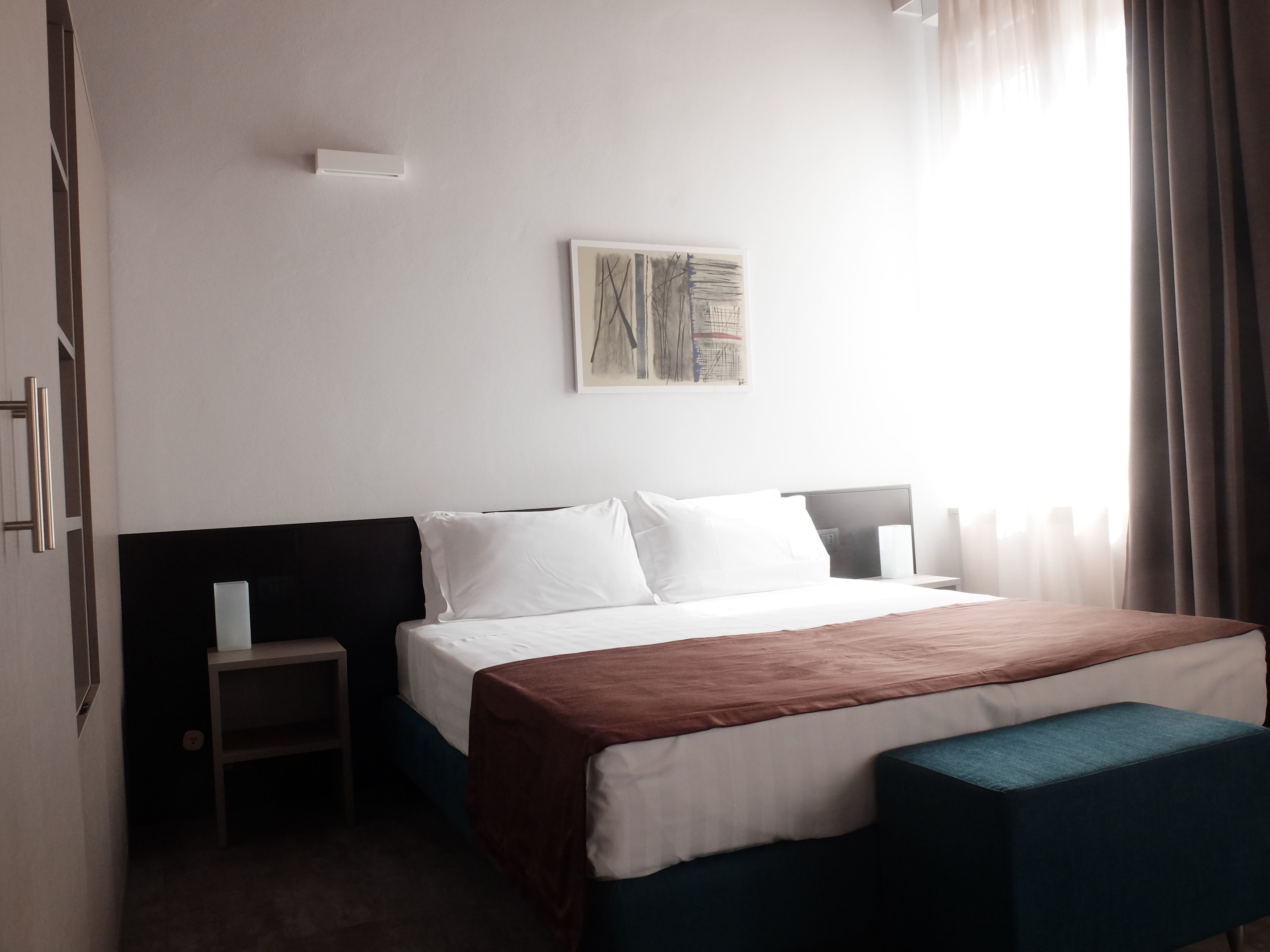 A hotel room with a bed, a bedside table, a bench, a wall-mounted artwork, and a window with curtains.