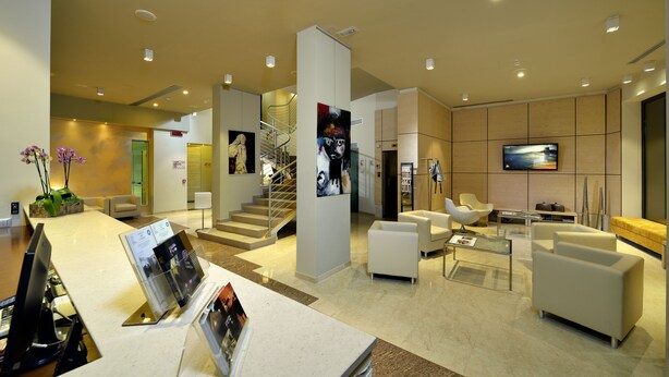 A modern hotel lobby with a reception desk, seating area, staircase, and a flat-screen TV. - Hotel Carlton (Ferrara)