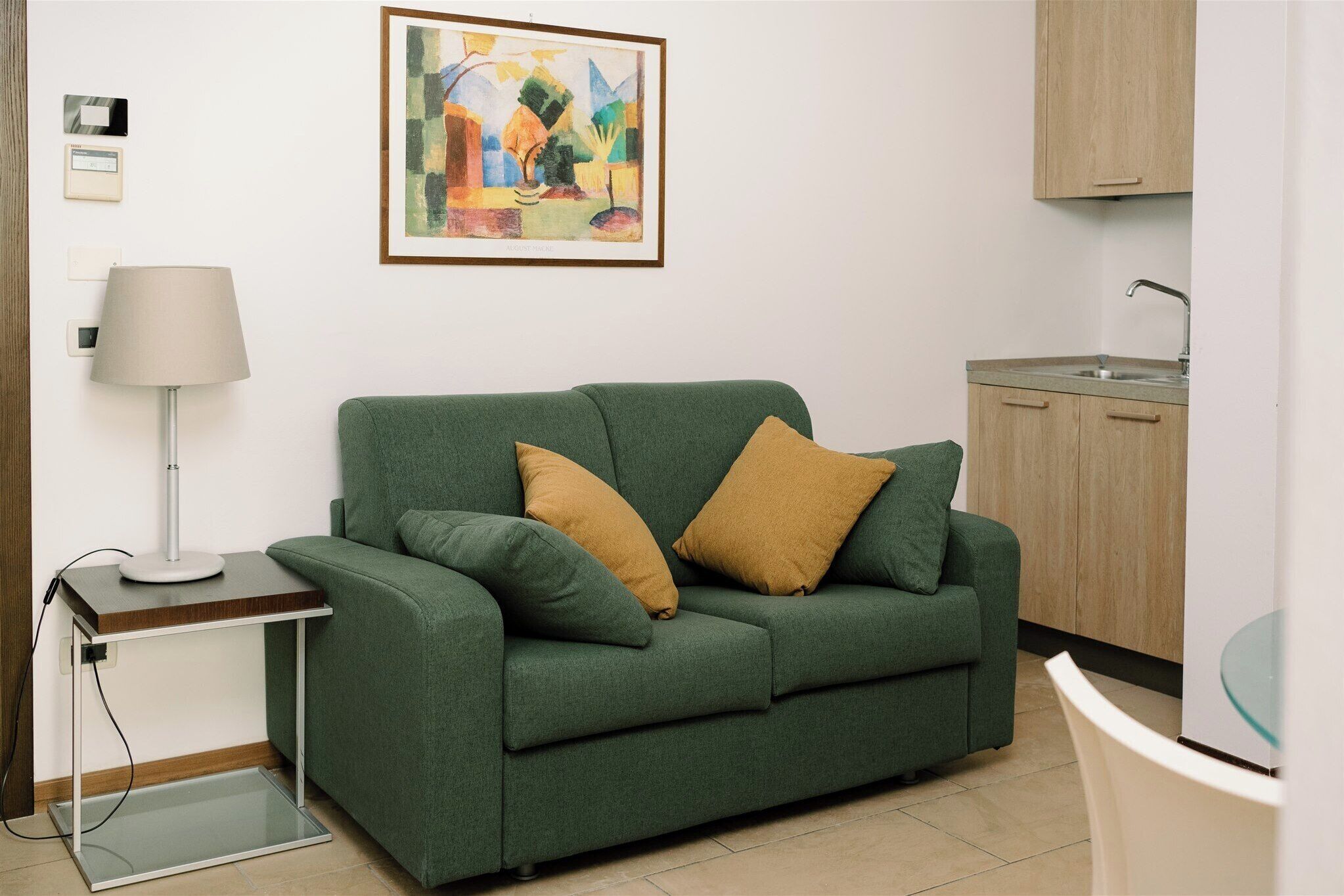 A green sofa with two pillows, a small table with a lamp, a framed abstract painting, a kitchen area with a sink and cabinets, and a glass table.