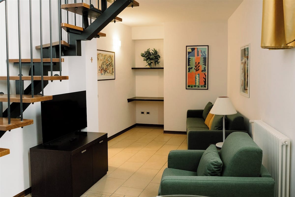 Una sala de estar con una escalera, un televisor de pantalla plana, un sofá verde, una lámpara y obras de arte en las paredes.