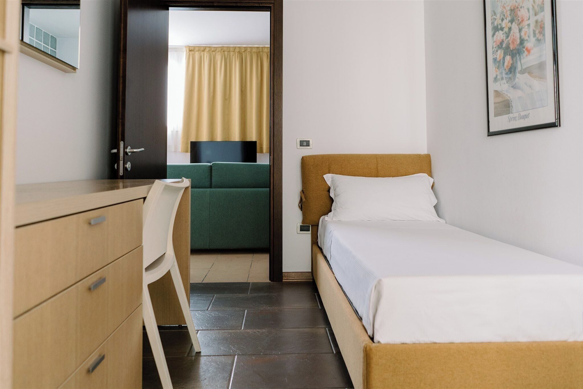Ein Hotelzimmer mit Bett, Schreibtisch, Stuhl und Blick auf ein weiteres Zimmer durch eine offene Tür.
