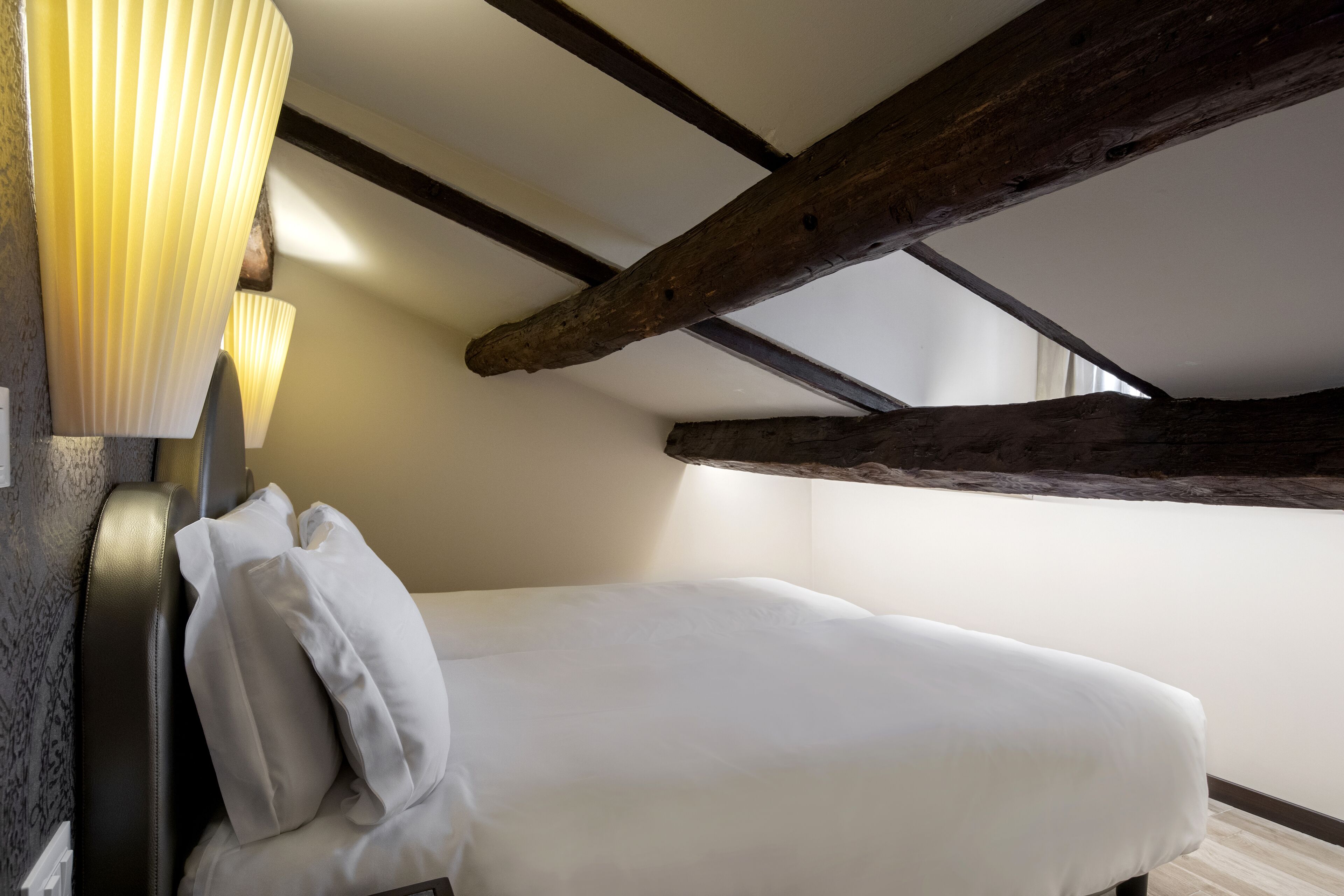 Ein Einzelbett mit weißer Bettwäsche in einem Raum mit freiliegenden Holzbalken und einer strukturierten Wand.