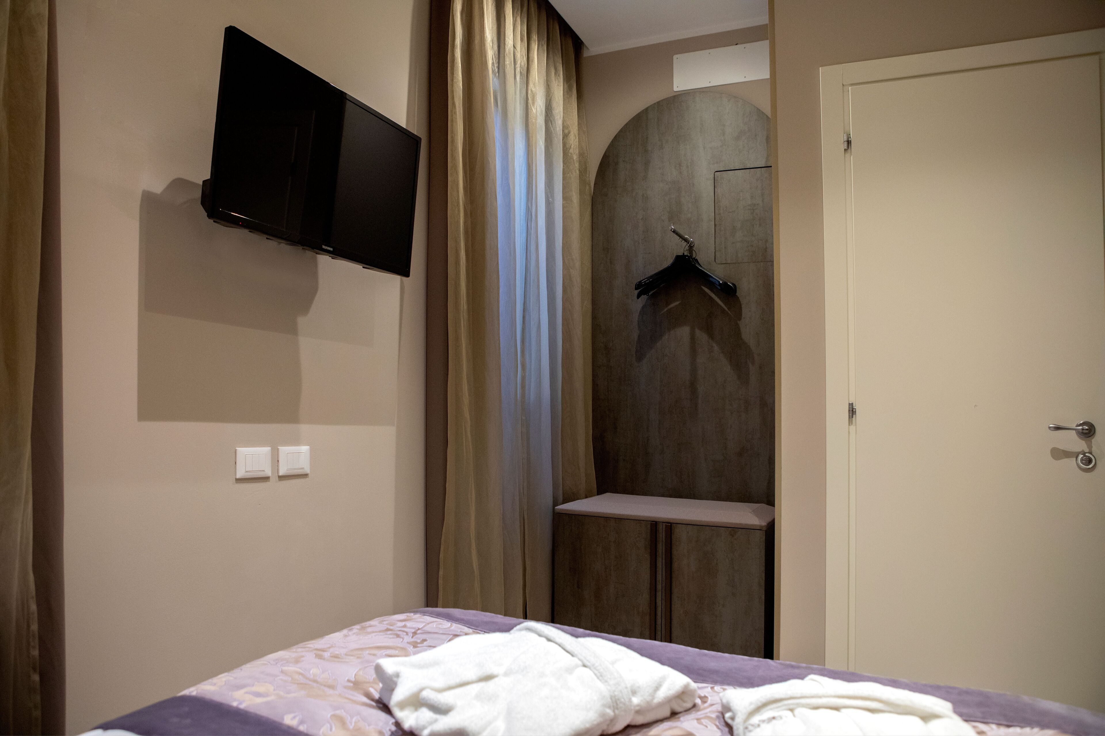 Una habitación de hotel con cama, televisor montado, perchero y puerta.