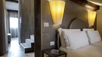 Una camera d'albergo con un letto, comodini e una rivista su uno dei comodini. Si vede una scala che conduce a un'altra stanza e una finestra con tende.