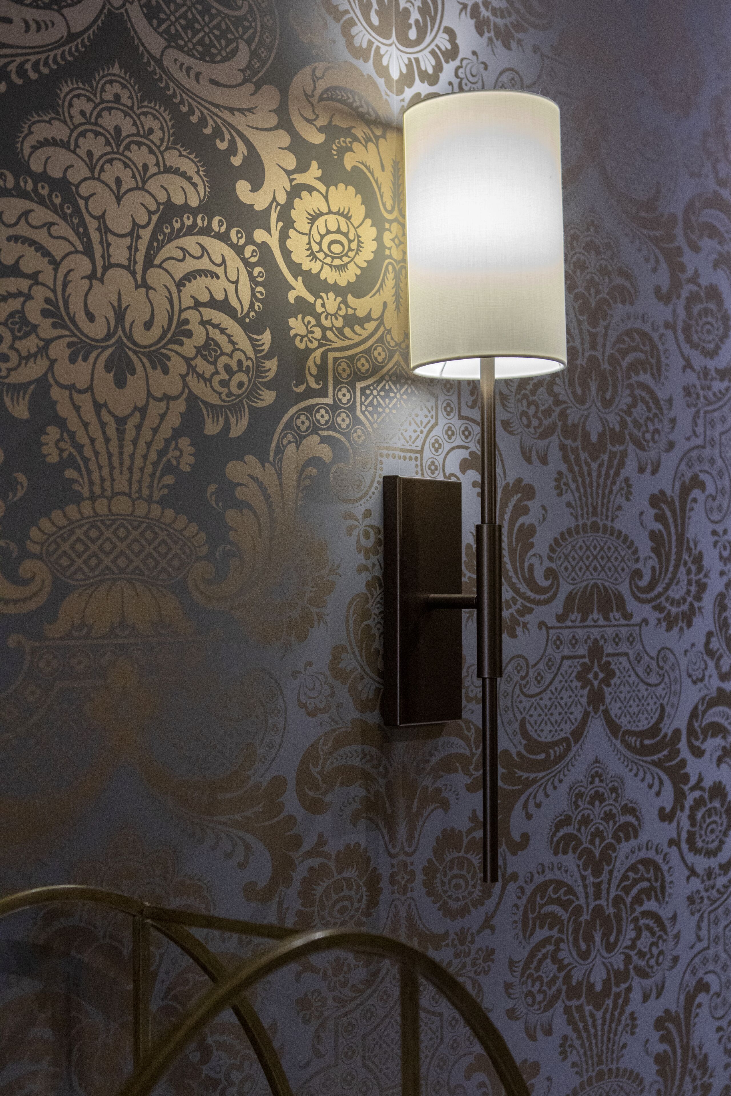 A wall-mounted lamp with a rectangular base and a cylindrical shade, positioned next to a wall with intricate floral and geometric patterns.