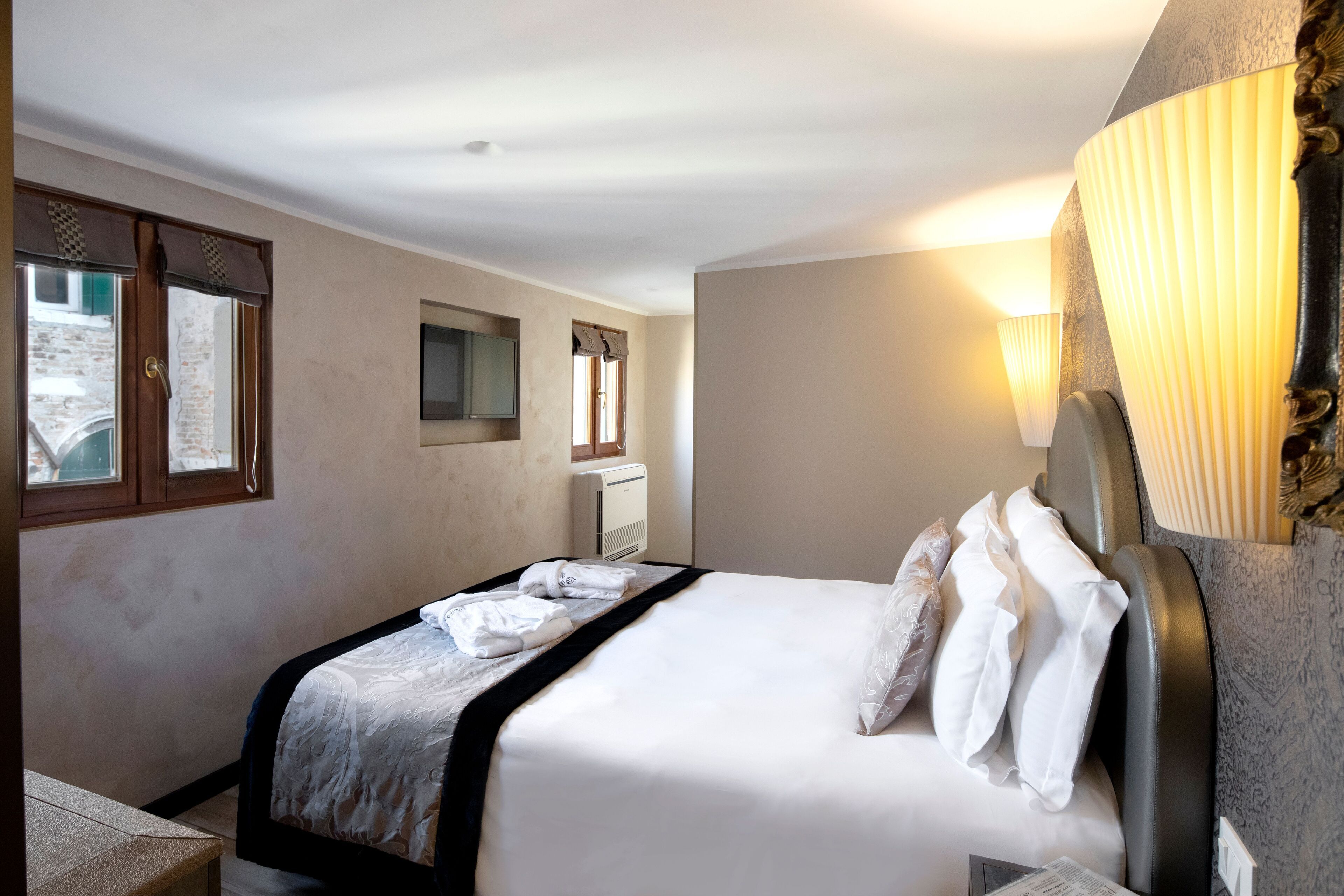 Una cama bien tendida con sábanas blancas, una mesita de noche, un televisor montado en la pared y una ventana con cortinas.