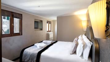 Una cama bien tendida con sábanas blancas, una mesita de noche, un televisor montado en la pared y una ventana con cortinas.