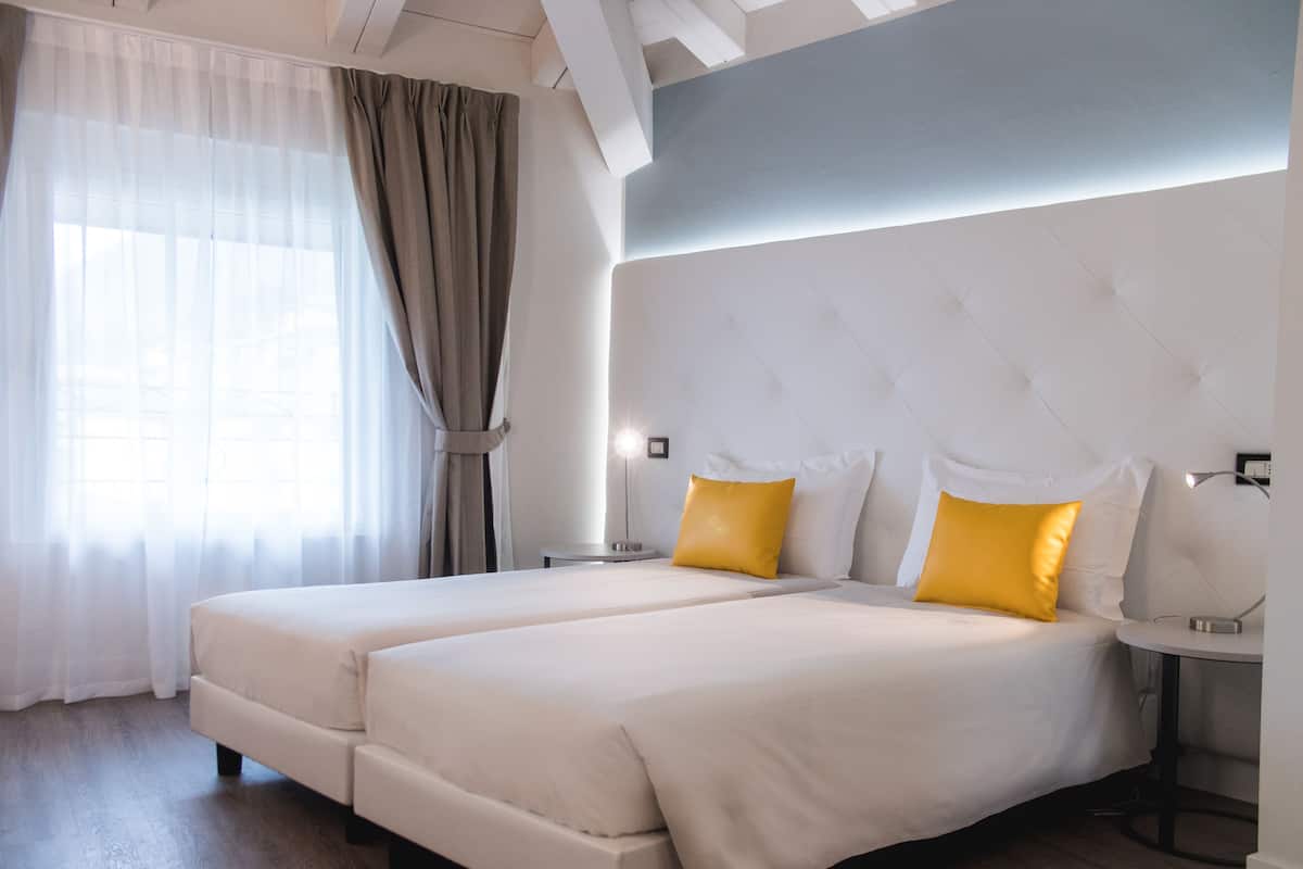 Une chambre d’hôtel moderne avec deux lits, une grande fenêtre avec des rideaux légers et un parquet en bois de couleur claire.
