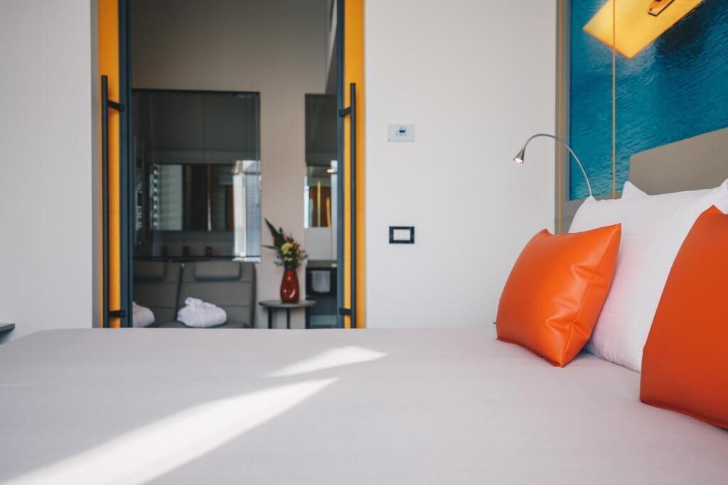A hotel room with a bed, orange pillows, a nightstand, a vase with flowers, and a painting on the wall.