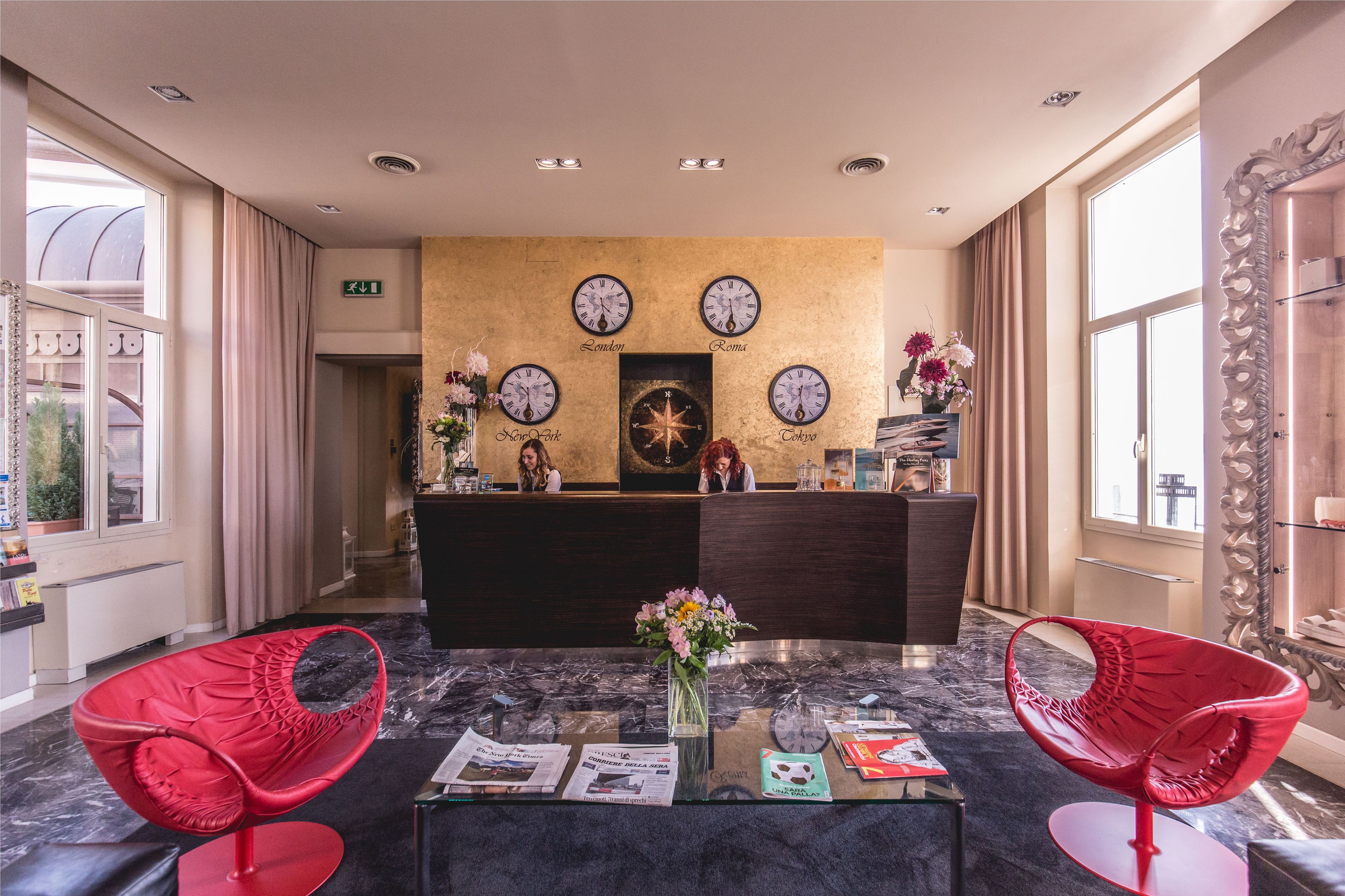 Un hall d’hôtel avec un comptoir d’accueil, deux fauteuils rouges, une table en marbre et des horloges indiquant différents noms de villes.