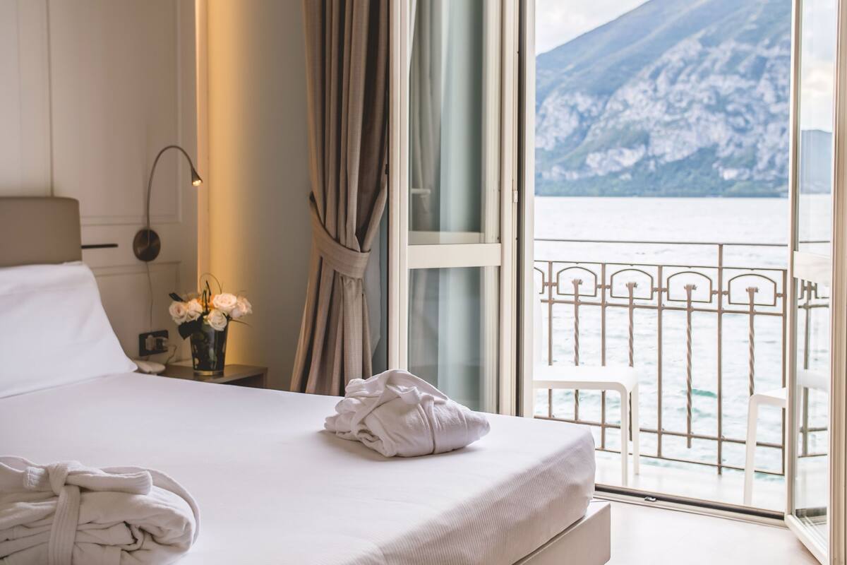 Une chambre d’hôtel avec un lit, une table de chevet avec une lampe, un vase de fleurs et une vue sur un paysage montagneux depuis un balcon.