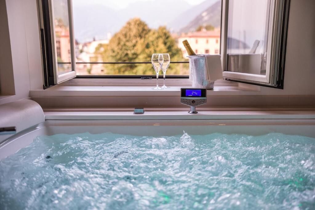 A hot tub with a digital thermometer, two glasses, and a champagne bottle on a windowsill with a view of buildings and trees.