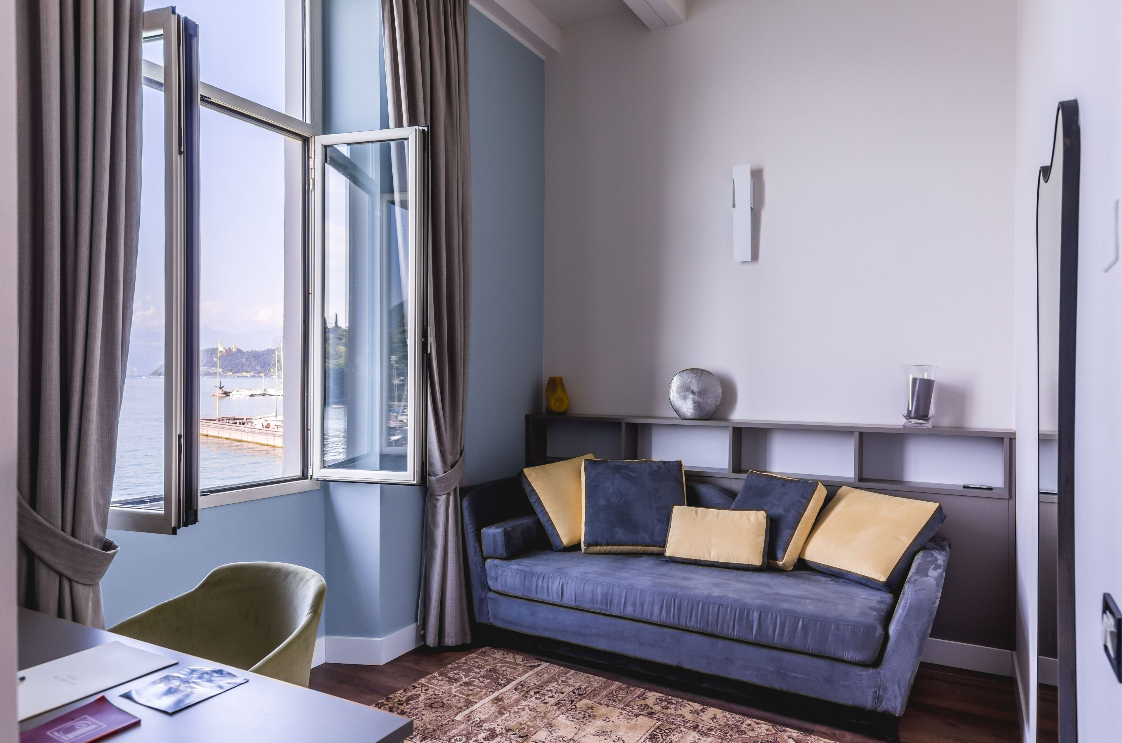 Ein modernes Wohnzimmer mit einem blauen Sofa, einem grünen Sessel und Blick auf ein Gewässer durch große Fenster.