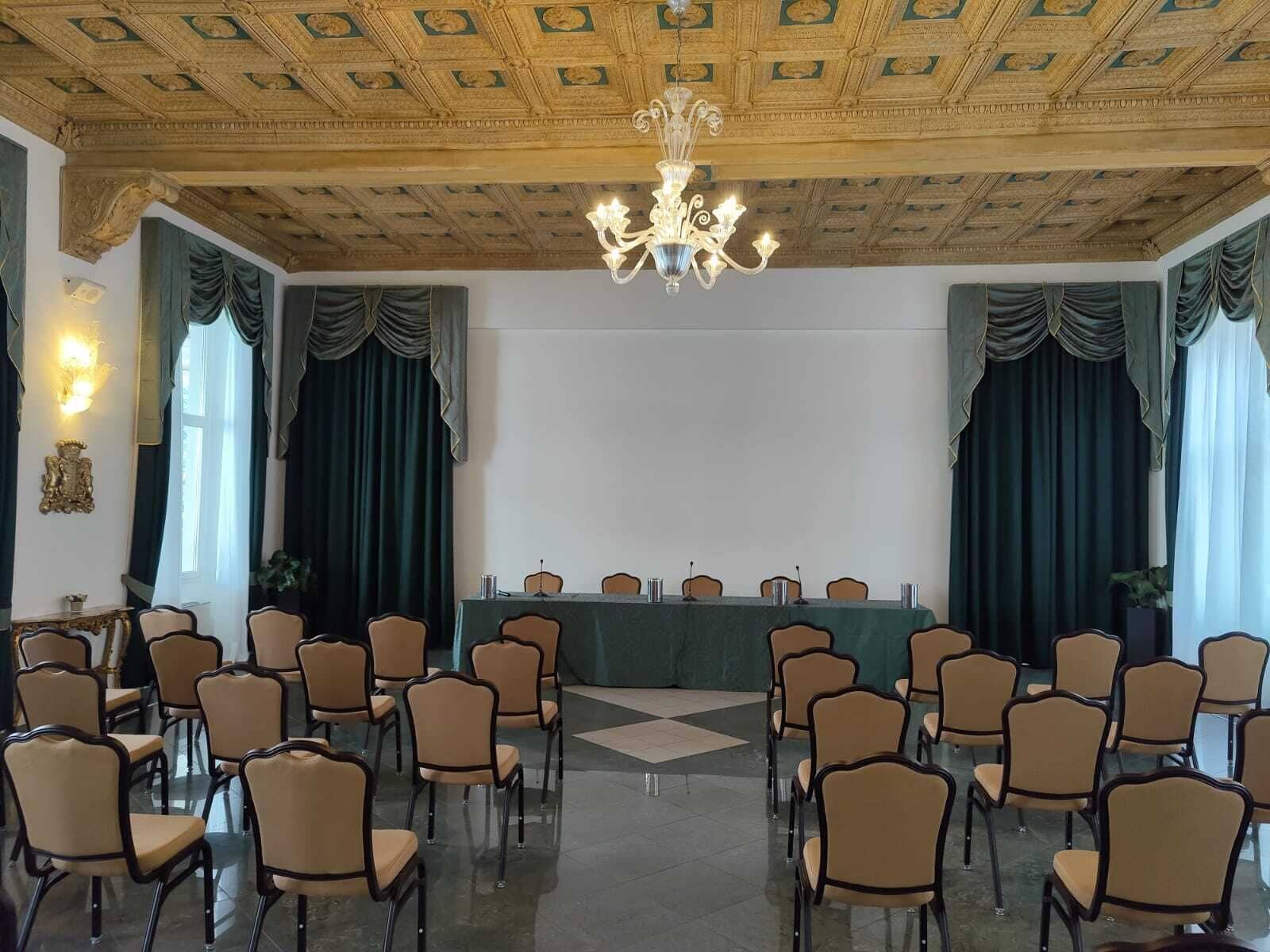 Un salón de conferencias con una larga mesa, sillas, una araña y cortinas con drapeado.