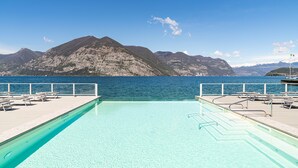 A swimming pool with clear blue water, surrounded by a wooden deck with lounge chairs and a view of mountains and the sea. - Hotel Araba Fenice (Iseo)
