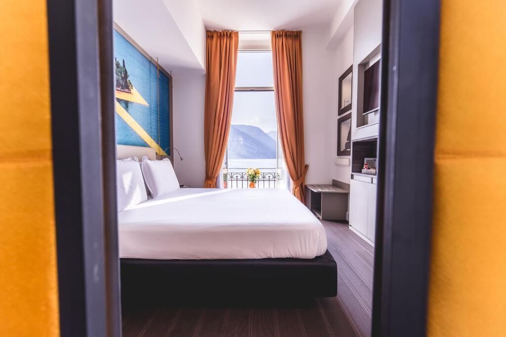 A hotel room with a bed, a desk, a chair, a window with curtains, and a painting on the wall.