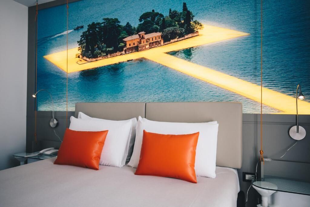 A hotel room with a bed, pillows, a nightstand, a lamp, and a large painting of a coastal scene.