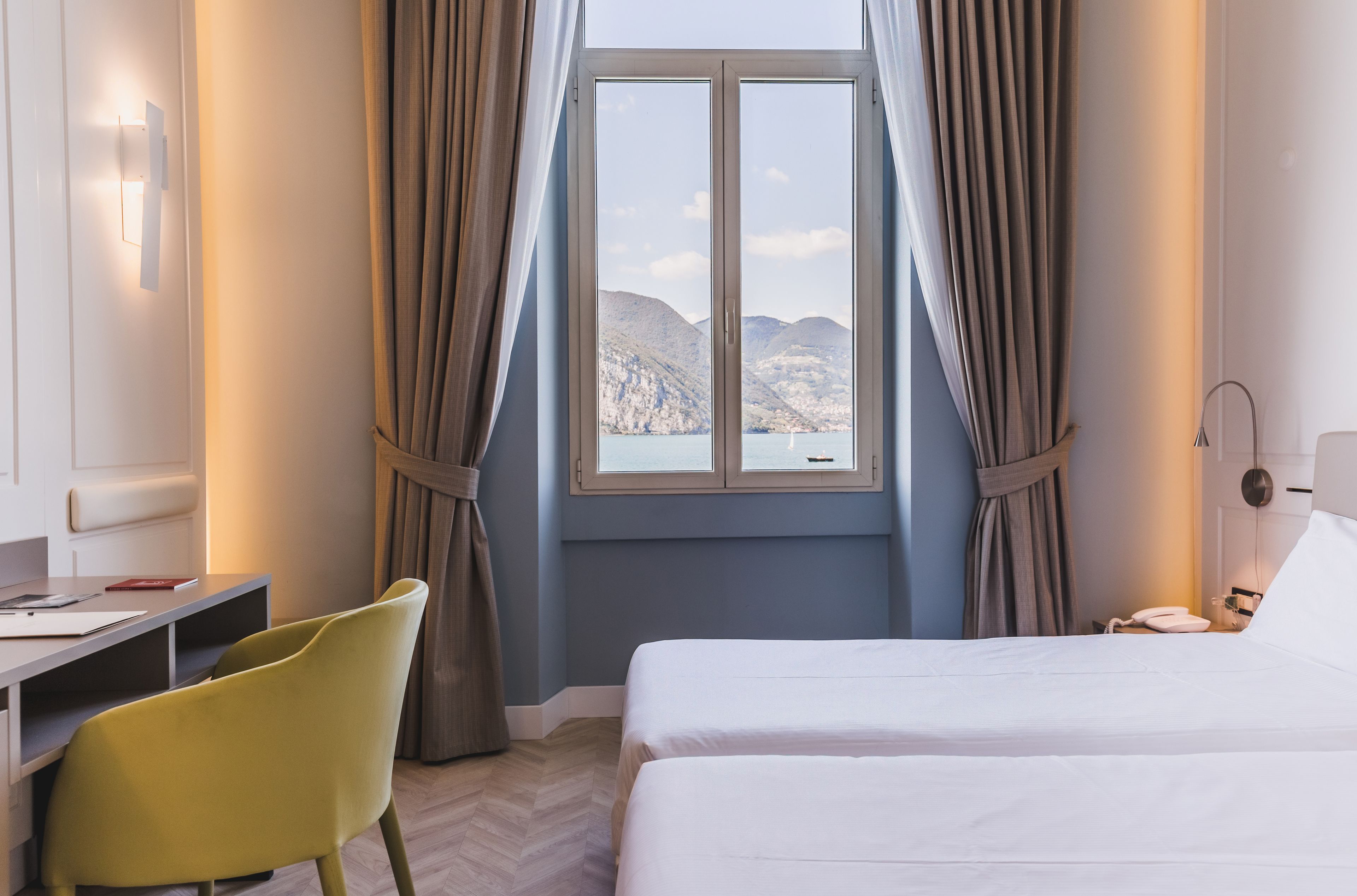 A hotel room with a bed, a desk, and a chair. There is a window with curtains, and a view of mountains outside.