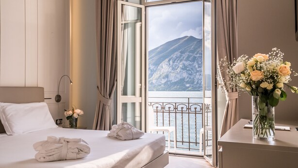 A hotel room with a bed, a nightstand with a lamp, a vase with flowers, and a view of mountains through a balcony. - Hotel Araba Fenice (Iseo)