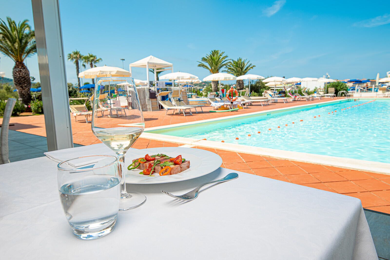 A dining table with a plate of food and a glass of water, overlooking a pool with lounge chairs and umbrellas.