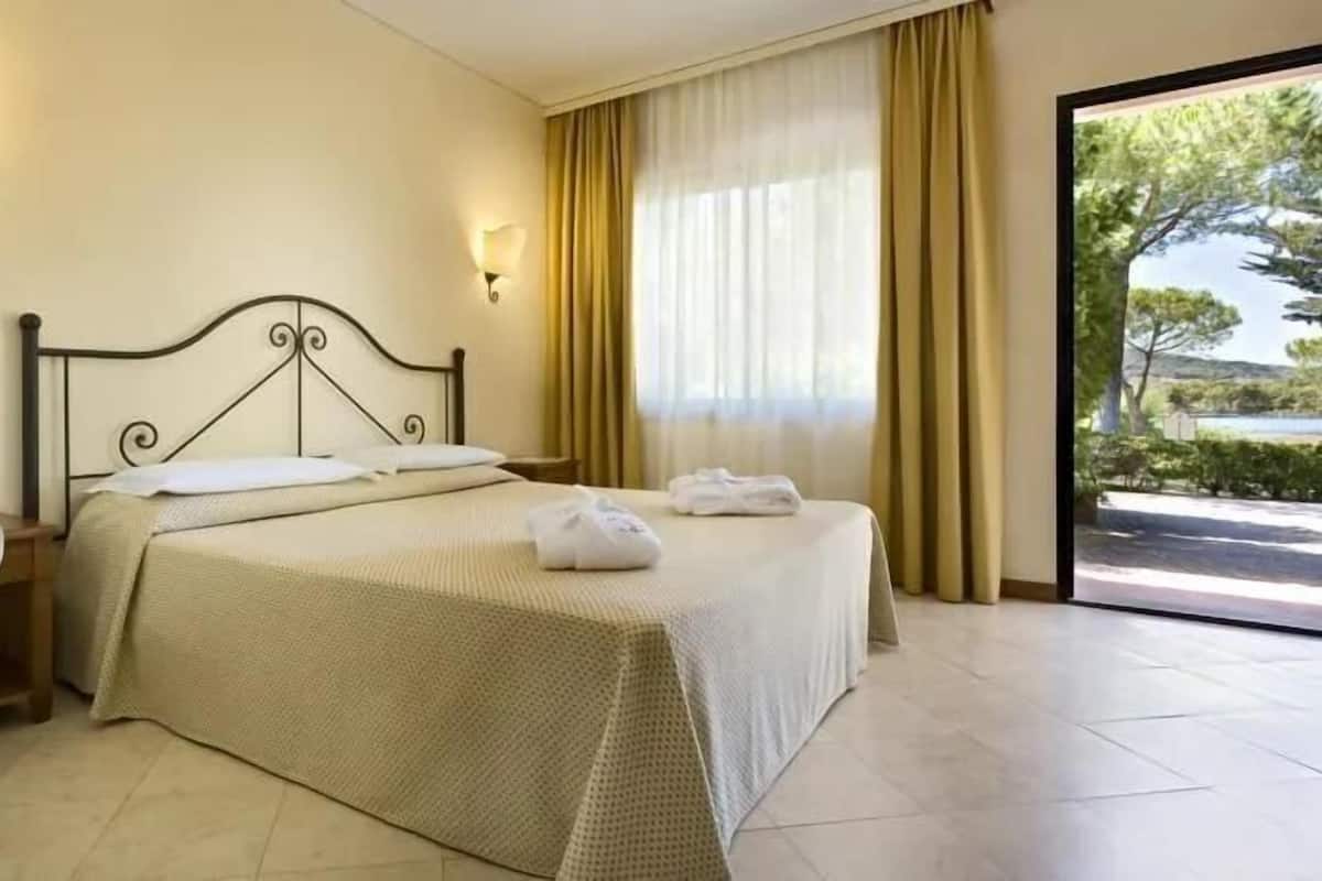 Ein Hotelzimmer mit einem Bett, einem Nachttisch mit Telefon und Blick auf Grün durch ein Fenster.