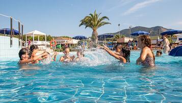 A swimming pool with people enjoying the water, surrounded by umbrellas and palm trees.