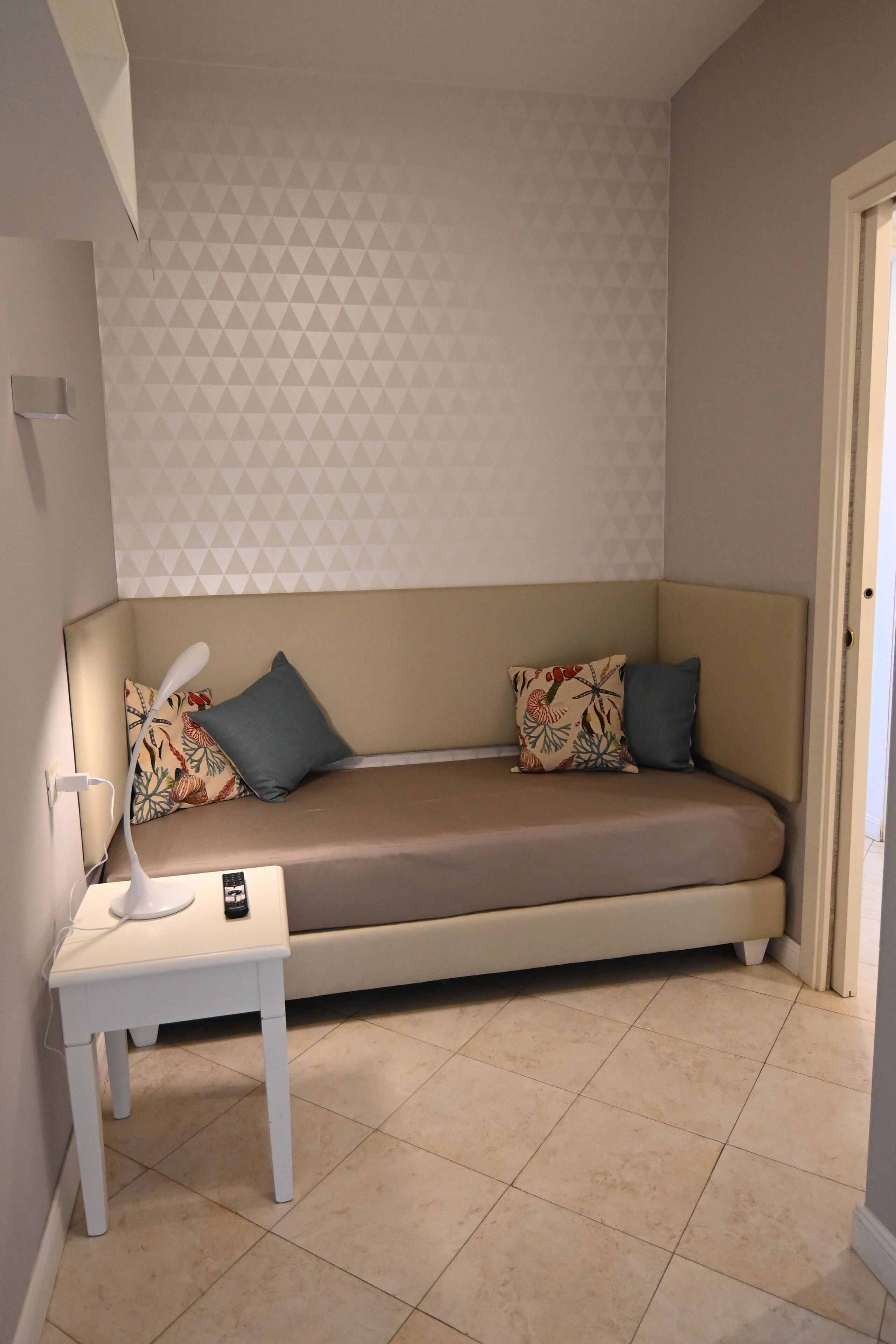A small, cozy room with a beige sofa, two pillows, a small white table with a lamp, and a patterned wall.
