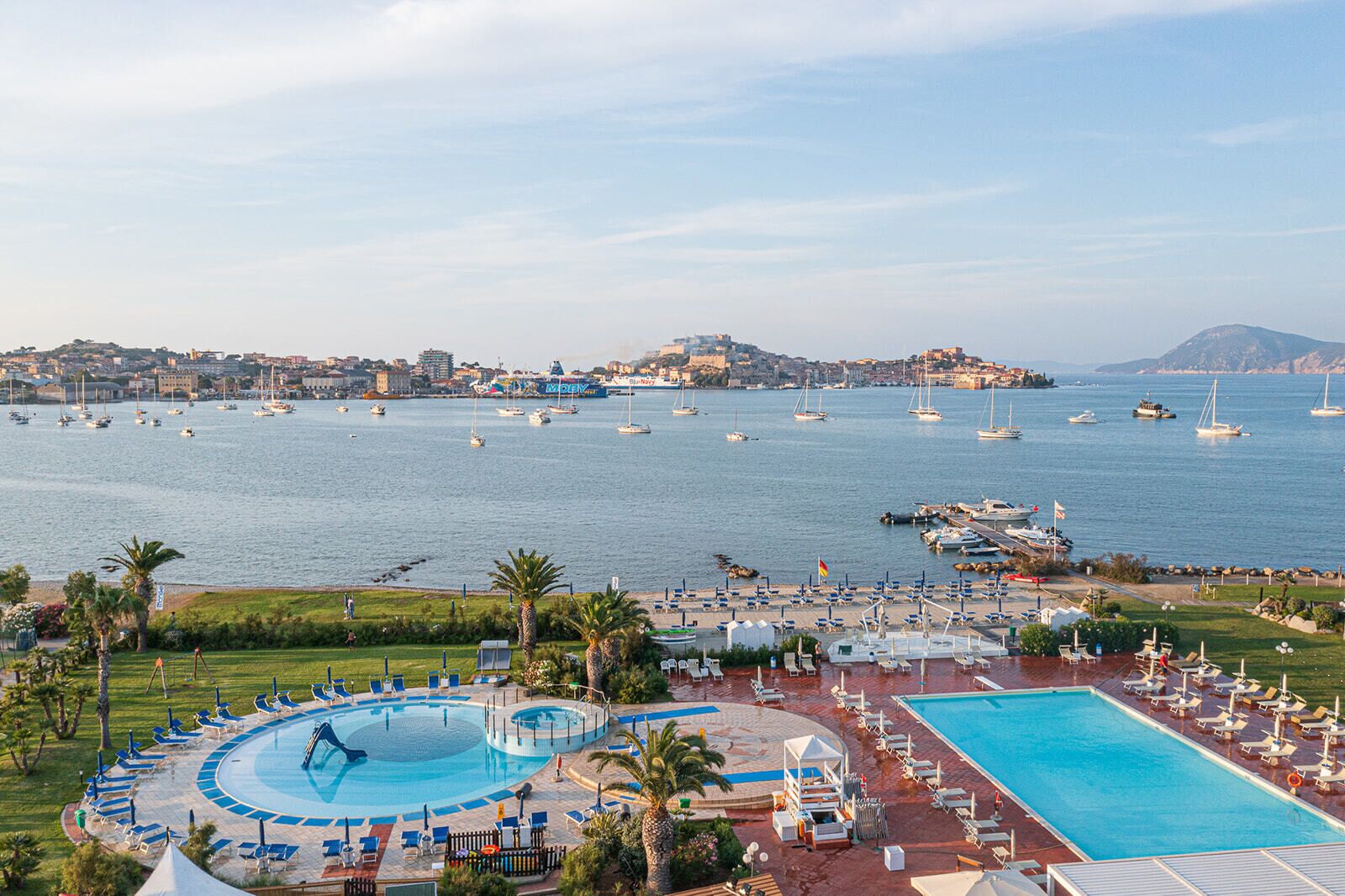 A resort with a large swimming pool, sun loungers, and a view of the sea with boats.