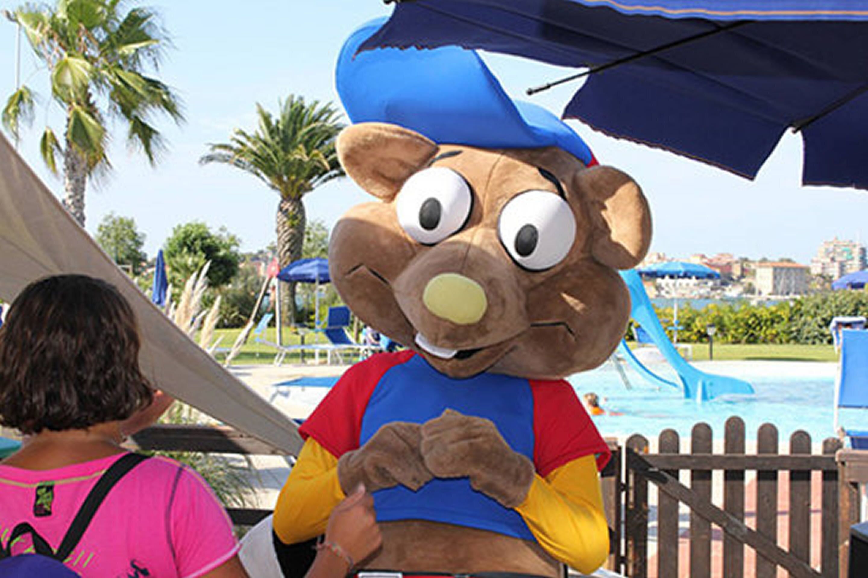 Un personaggio con un costume da mascotte in piedi vicino a una piscina, con palme e uno scivolo d'acqua sullo sfondo.