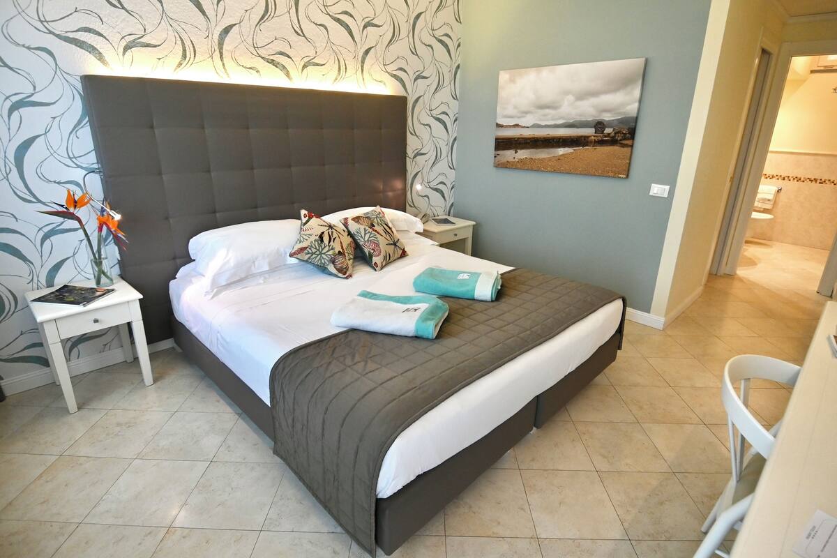 Ein Hotelzimmer mit einem großen Bett, einem Nachttisch, einer Wandlampe, einem Gemälde und einem durch eine offene Tür einsehbaren Badezimmer.