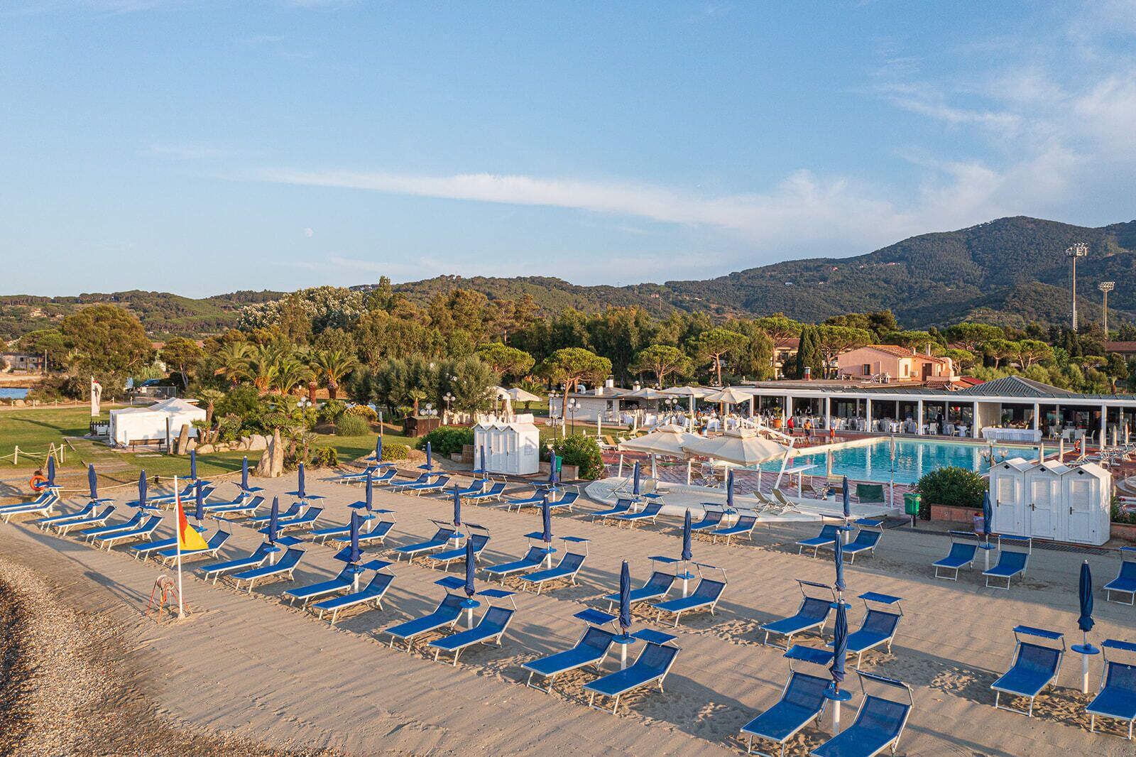 A beachside resort with multiple sun loungers and umbrellas, a swimming pool, and a view of the surrounding hills.