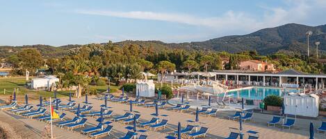 A beachside resort with multiple sun loungers and umbrellas, a swimming pool, and a view of the surrounding hills.