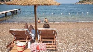 Een strand met twee houten ligstoelen onder een strooien parasol, een kiezelstrand en een rustige zee met boten in de verte.