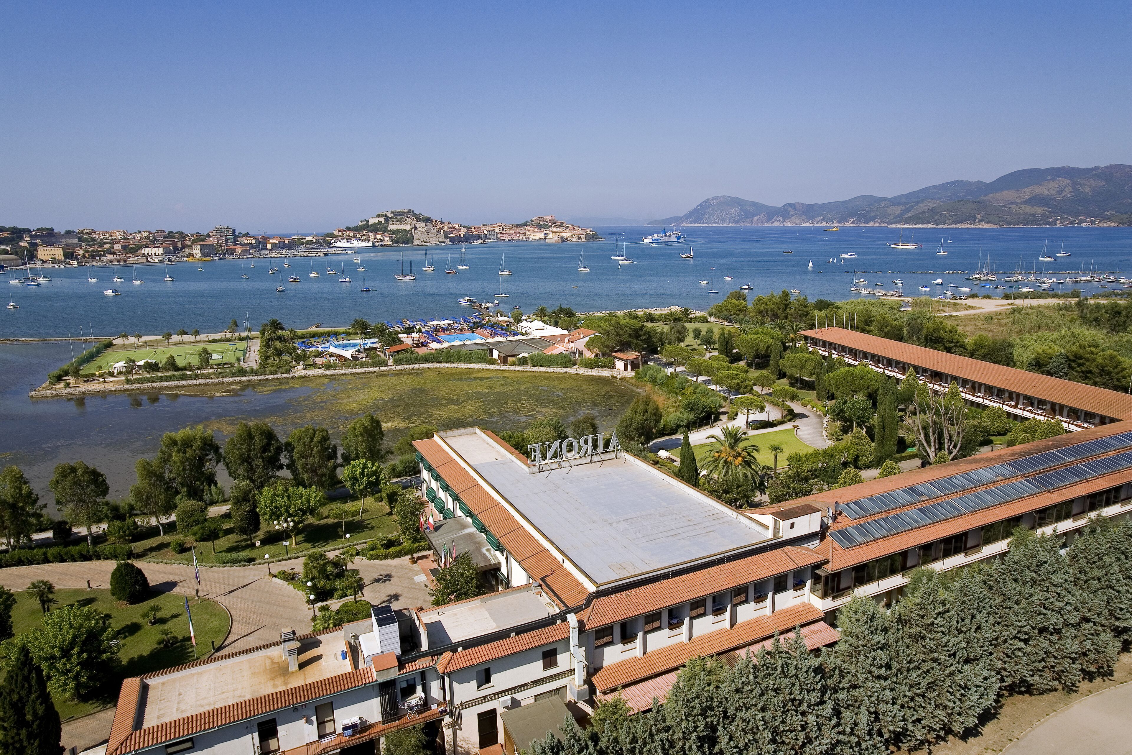 Un edificio costero con techo plano y paneles solares, rodeado de vegetación y un puerto deportivo con barcos.