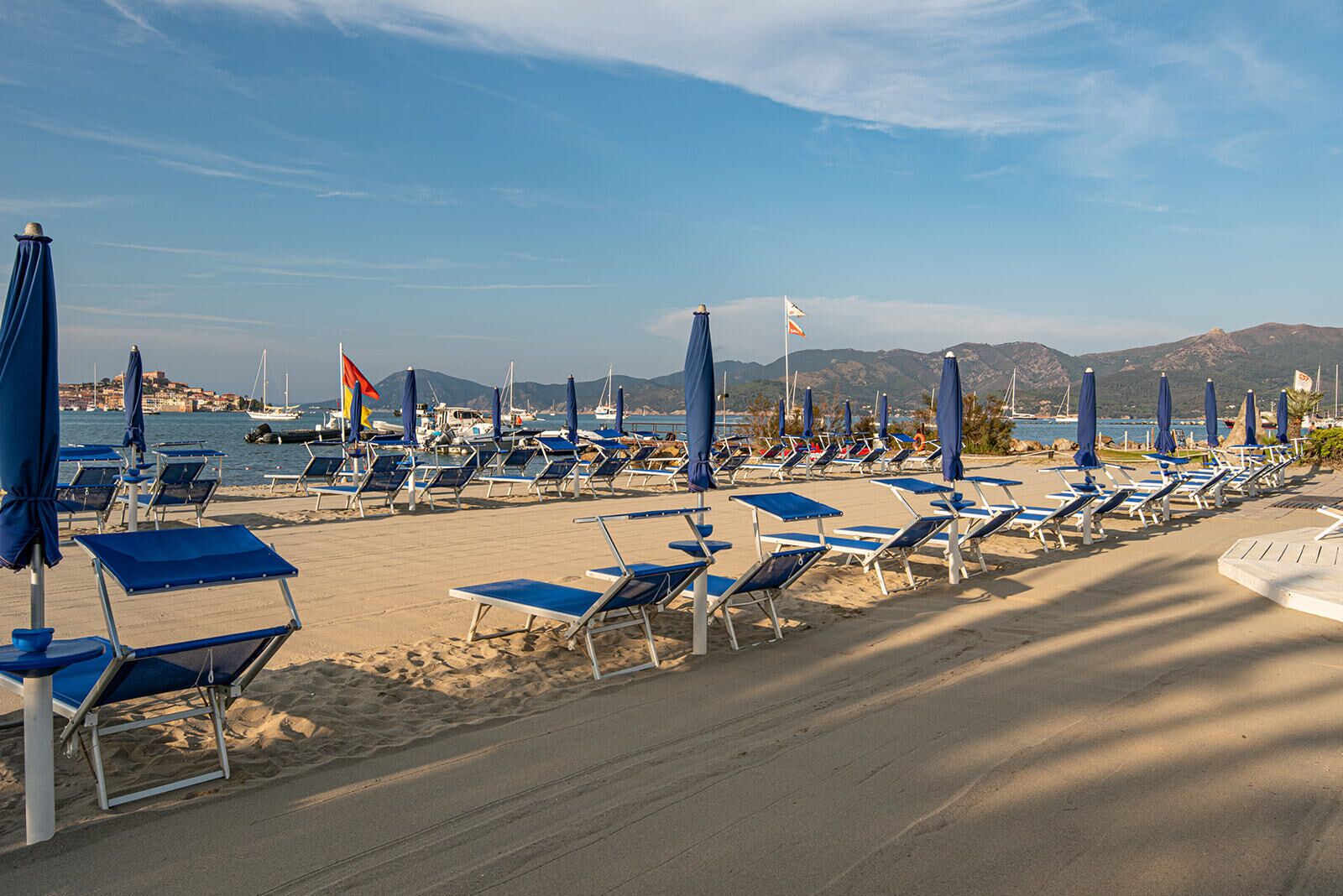 A beach with rows of sun loungers and umbrellas, a clear sky, and boats in the background.