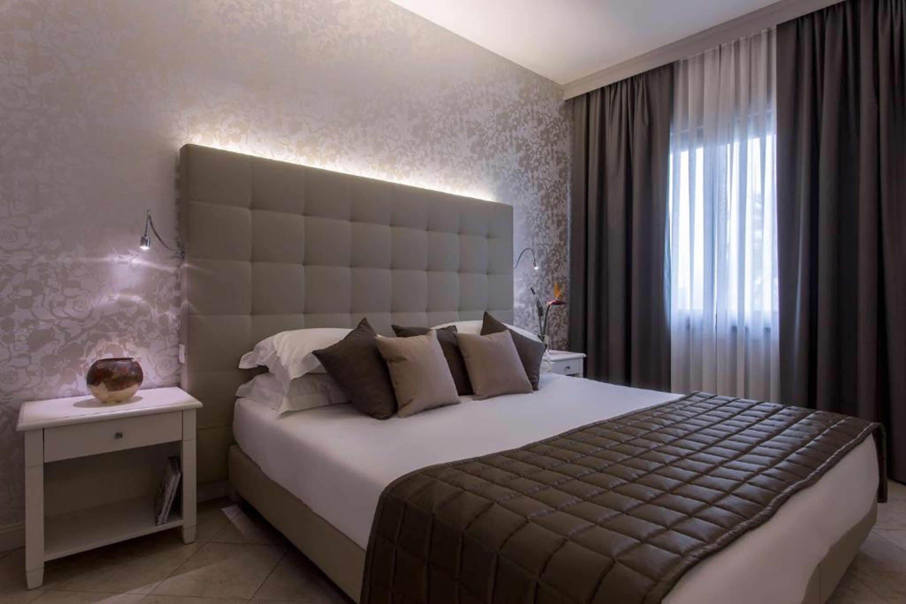 A modern bedroom with a tufted headboard, a large bed with a brown quilt, a bedside table with a vase, and a window with curtains.