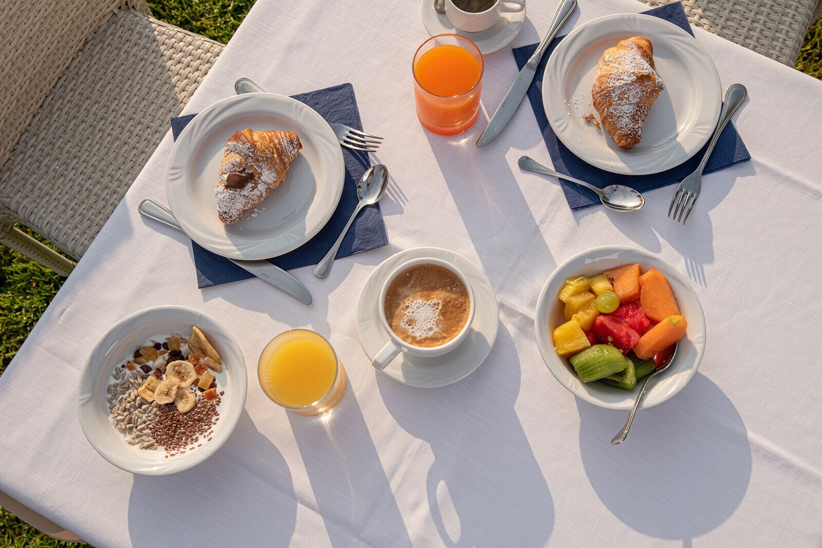 A table set outdoors with breakfast items including croissants, fruit, cereal, and coffee.