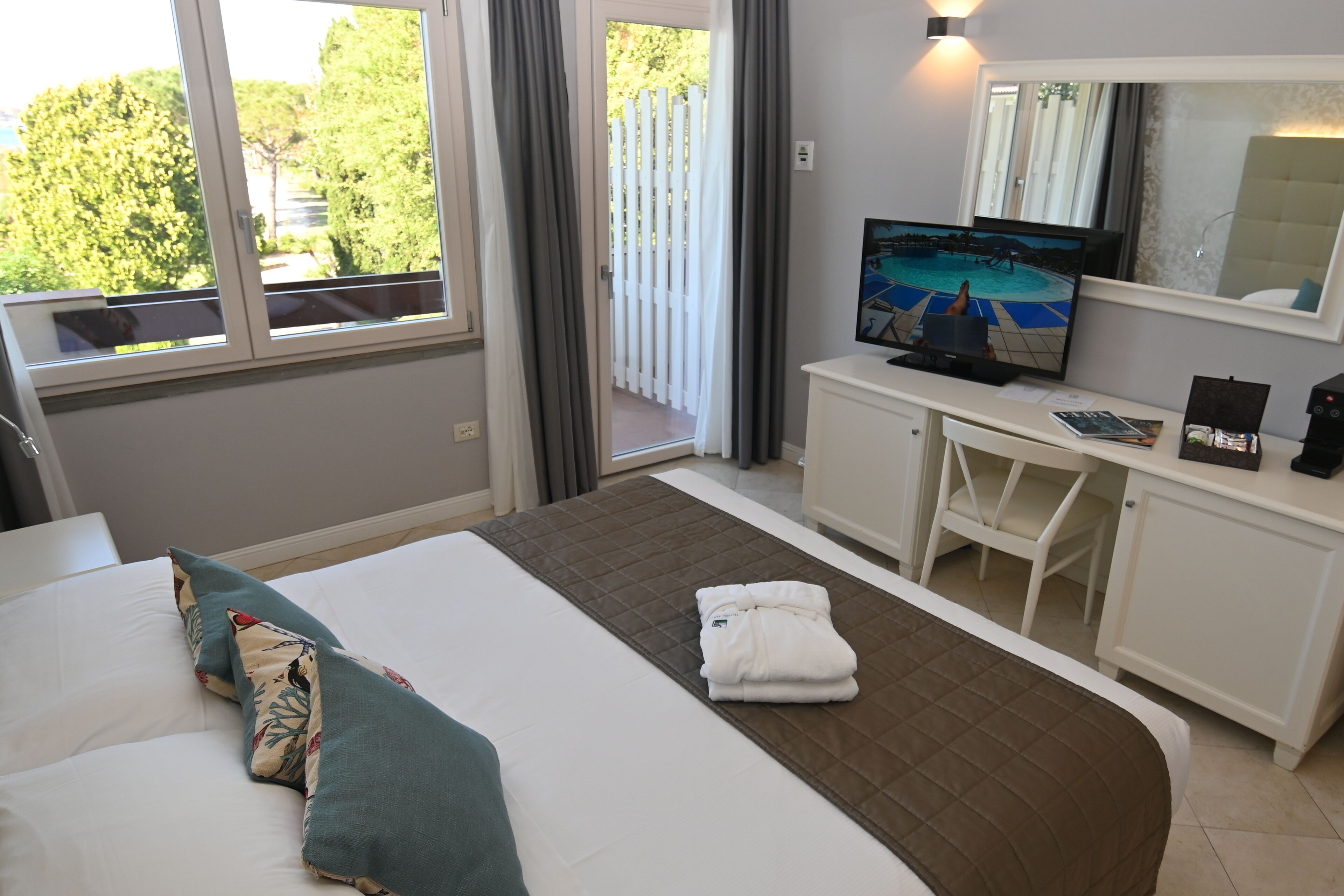 A hotel room with a bed, a desk, a television, and a view of trees and a pool.