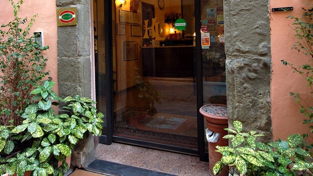 Entrada de um hotel com um arco, um tapete na porta com o nome do hotel e vasos de plantas de ambos os lados.
