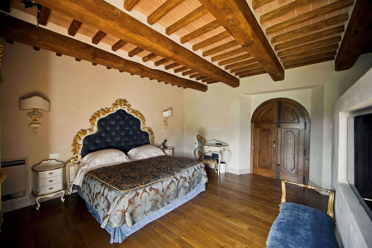 A bedroom with a large bed, wooden ceiling beams, a fireplace, and a wooden wardrobe.