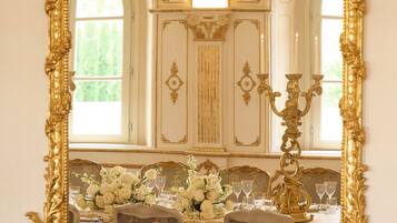 A grand mirror with an ornate gold frame, reflecting a room with a table set for a meal, a candelabra, and a window with natural light.