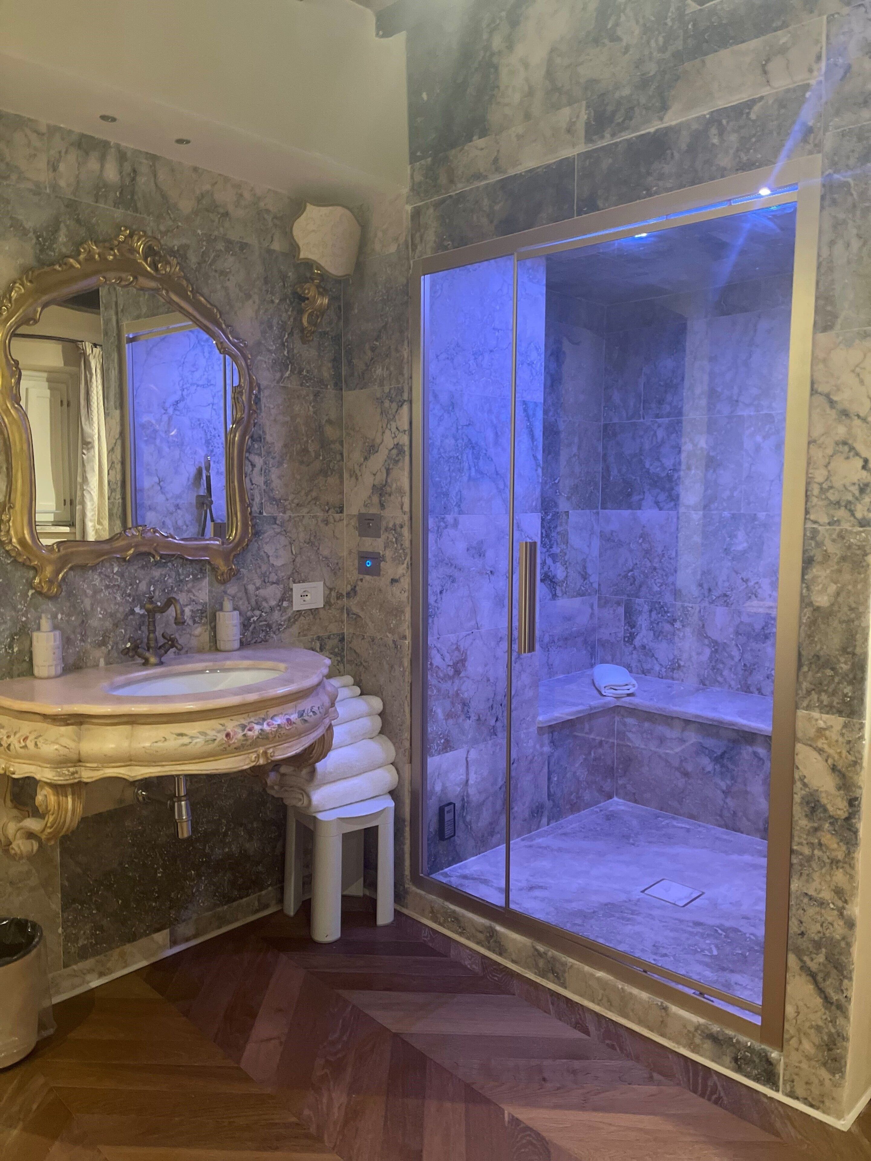 Ein Badezimmer mit Marmor-Waschtisch, einem großen, verzierten Spiegel und einer gläsernen Duschkabine.