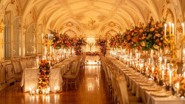 A grand banquet hall with ornate ceiling, long tables set for a formal event, and elaborate floral centerpieces.
